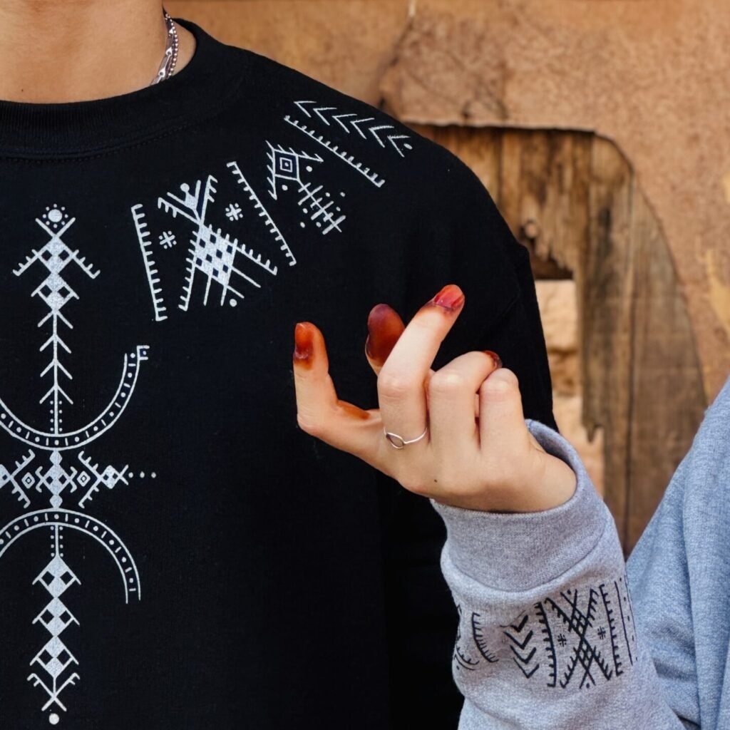 Black sweatshirt with white Amazigh geometric motifs beside a gray patterned sleeve