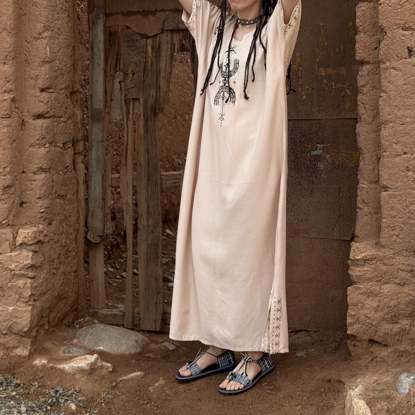Cream Moroccan Amazigh dress with black geometric embroidery worn in a traditional earthen doorway
