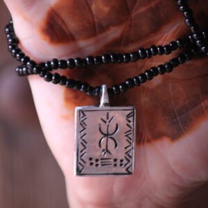 Pendentif amazigh en métal plaqué argent avec motif gravé, monté sur un collier de perles noires.
