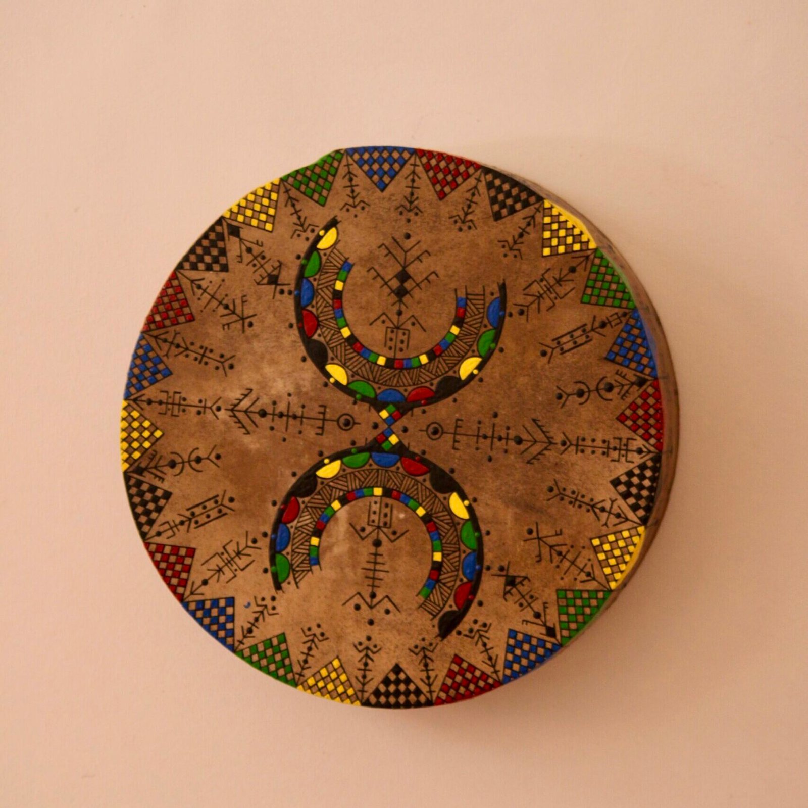 Bendir peint à la main par Muha Tawargit, décoré de motifs amazighs noirs et colorés, avec une main ornée de henné au premier plan.