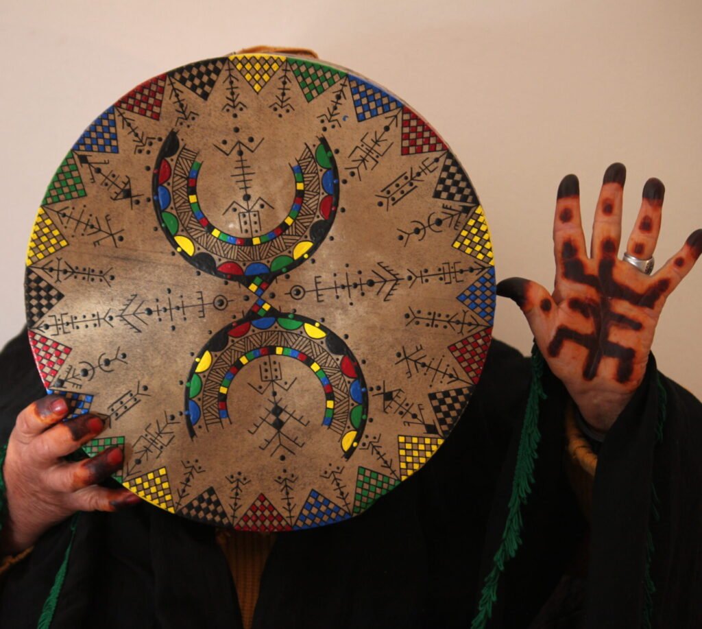 Bendir peint à la main par Muha Tawargit, décoré de motifs amazighs noirs et colorés, avec une main ornée de henné au premier plan.