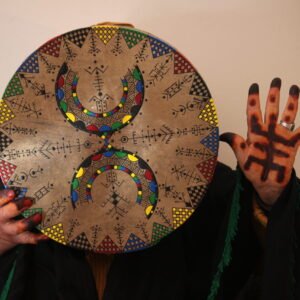 Bendir peint à la main par Muha Tawargit, décoré de motifs amazighs noirs et colorés, avec une main ornée de henné au premier plan.