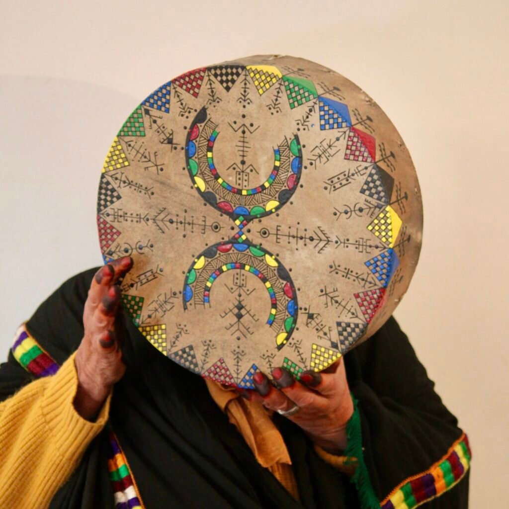 Bendir peint à la main par Muha Tawargit, décoré de motifs amazighs noirs et colorés, avec une main ornée de henné au premier plan.