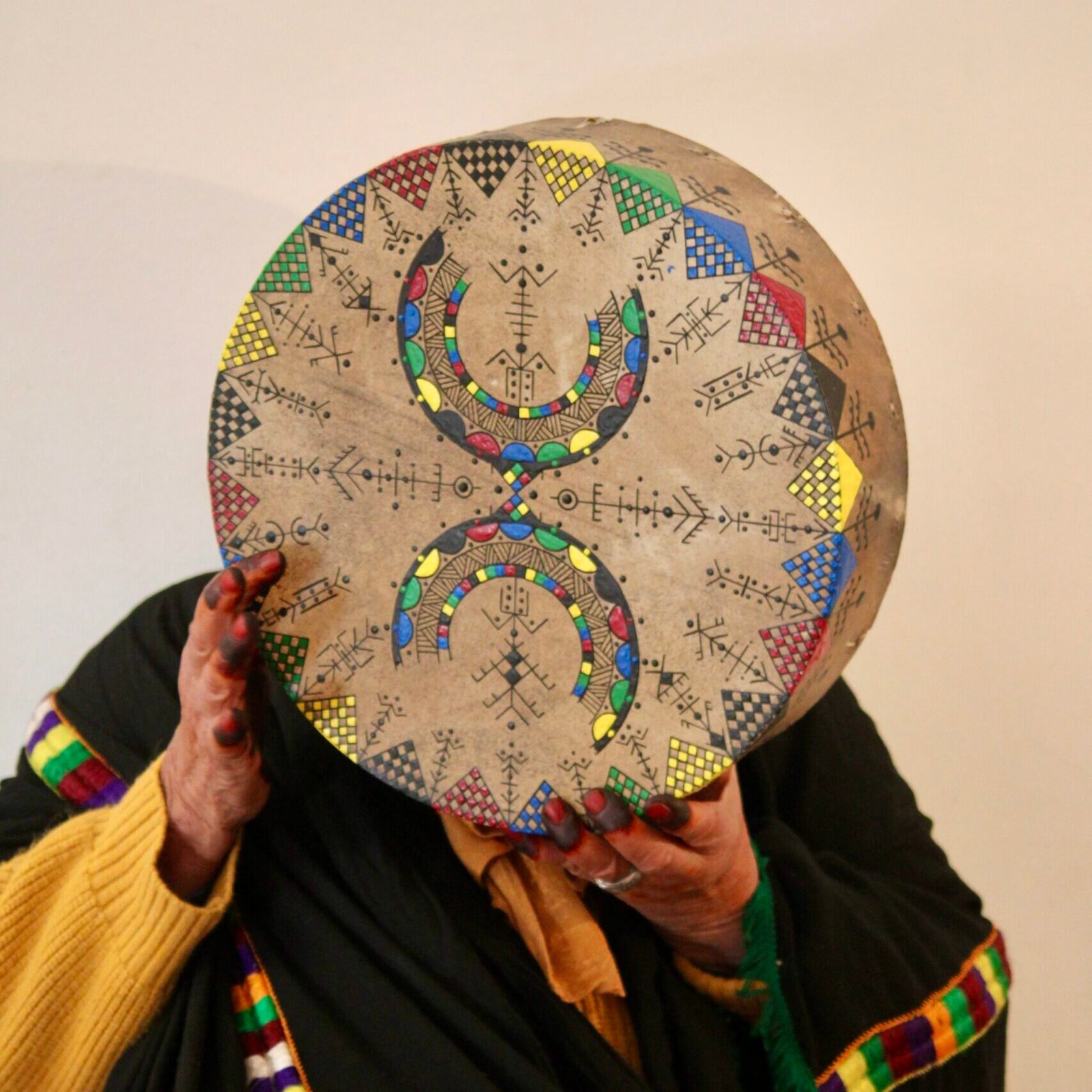 Bendir peint à la main par Muha Tawargit, décoré de motifs amazighs noirs et colorés, avec une main ornée de henné au premier plan.