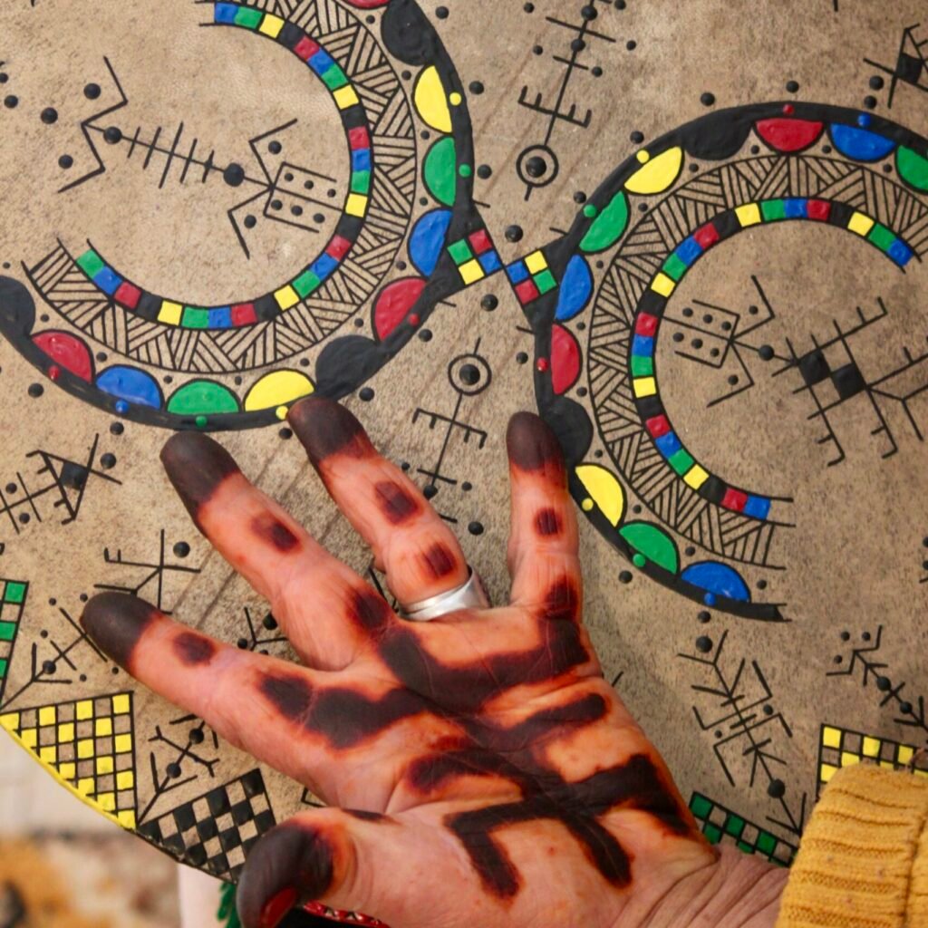 Bendir peint à la main par Muha Tawargit, décoré de motifs amazighs noirs et colorés, avec une main ornée de henné au premier plan.