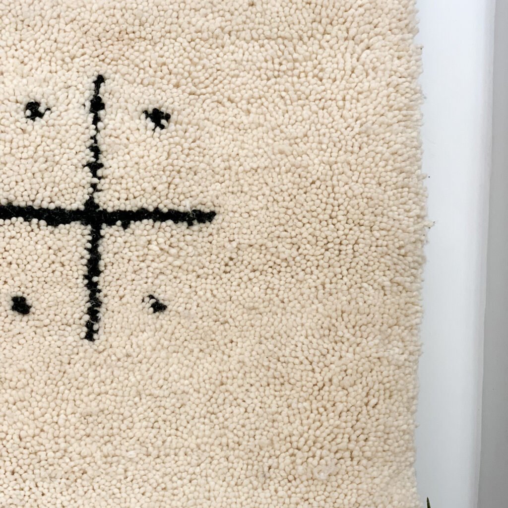 Tapis artisanal écru conçu par Afusart, tissé à la main par Malika, avec motifs amazighs noirs et touches colorées.