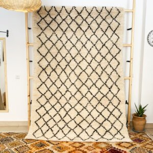 Tapis artisanal écru conçu par Afusart, tissé à la main par Malika, avec motifs amazighs noirs et touches colorées.