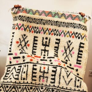 Tapis artisanal écru conçu par Afusart, tissé à la main par Malika, avec motifs amazighs noirs et touches colorées.