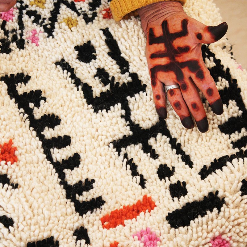 Tapis artisanal écru conçu par Afusart, tissé à la main par Malika, avec motifs amazighs noirs et touches colorées.